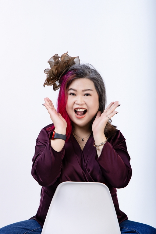 Joyful personal branding portrait of Aunty HR, a Malaysian HR influencer, wearing a burgundy blouse with pink hair highlights and a floral accessory, smiling enthusiastically against a crisp white studio background.